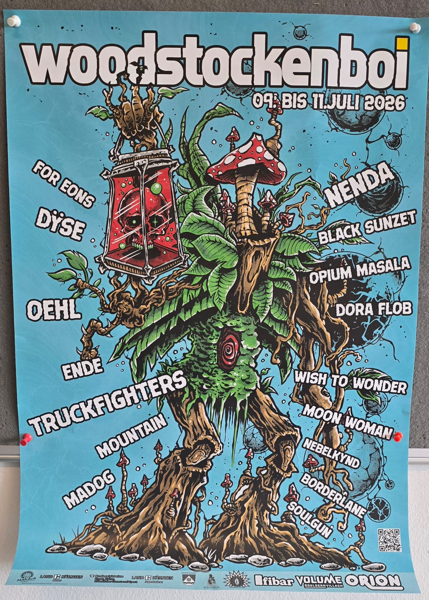 Buntes Festivalplakat für „woodstockenboi“ vom 9. bis 11. Juli 2026. Im Zentrum steht eine fantasievolle, monsterartige Baumfigur mit Wurzeln, Pilzen, Pflanzen und einem roten Auge. Um die Figur herum sind die Namen verschiedener Bands und Künstler angeordnet, darunter Nenda, Black Sunzet, Truckfighters und Moon Woman. Das Design ist im Comic- und Psychedelic-Stil gehalten, mit kräftigen Farben und vielen Details auf hellblauem Hintergrund.