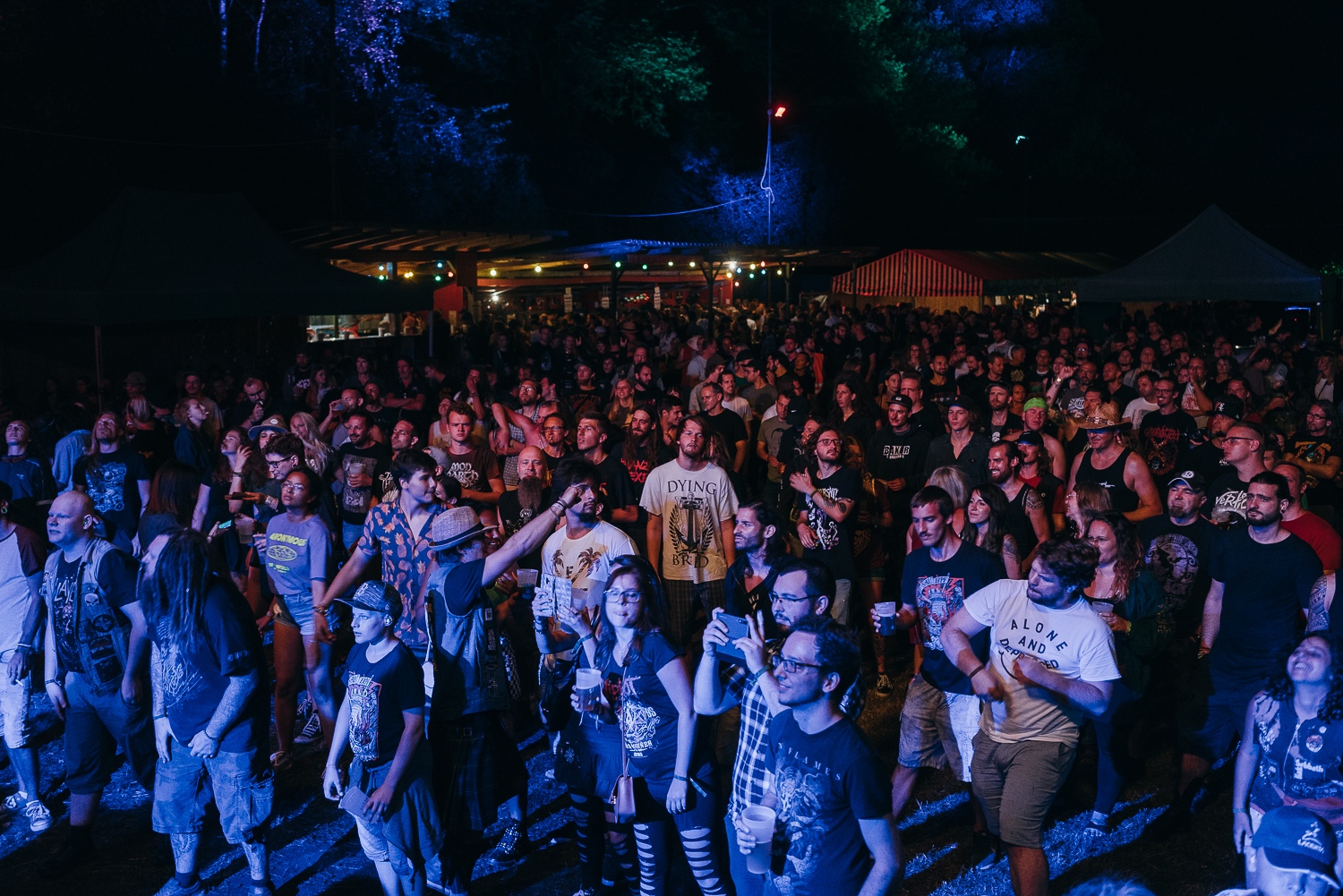 Große Menschenmenge bei einem nächtlichen Konzert Die Besucher stehen dicht gedrängt vor der Bühne, viele tragen dunkle Bandshirts und halten Getränke in der Hand. Bunte Lichter und beleuchtete Stände sorgen für Festivalstimmung.