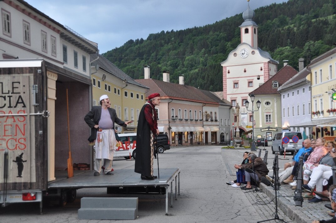 Auf einem mobilen Theaterwagen am Hauptplatz in der Altstadt von Gmünd stehen zwei Schauspieler in historischen Kostümen. Im Hintergrund sind pastellfarbene Häuser, eine Kirche mit Turm und bewaldete Hügel zu sehen. Rechts sitzt ein kleines Publikum auf Stühlen und verfolgt die Aufführung unter freiem Himmel.