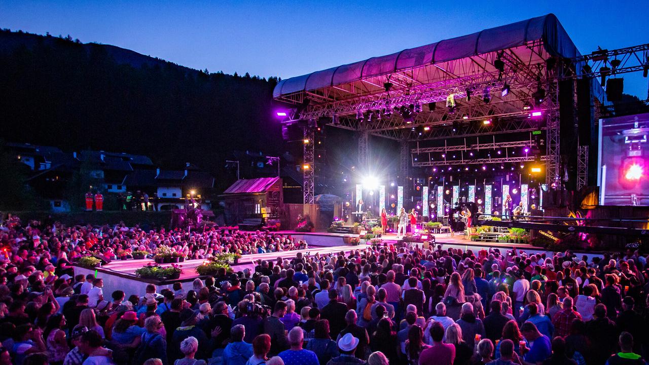 Große Open-Air-Veranstaltung „Wenn die Musie spielt“ bei Abendstimmung in den Bergen. Eine beleuchtete Konzertbühne mit Sängerinnen, Musikern und farbigen Lichteffekten steht vor einem großen Publikum. Zahlreiche Besucherinnen und Besucher verfolgen das Konzert vor der Bühne, umgeben von einer alpinen Landschaft und dunklen Berghängen im Hintergrund.