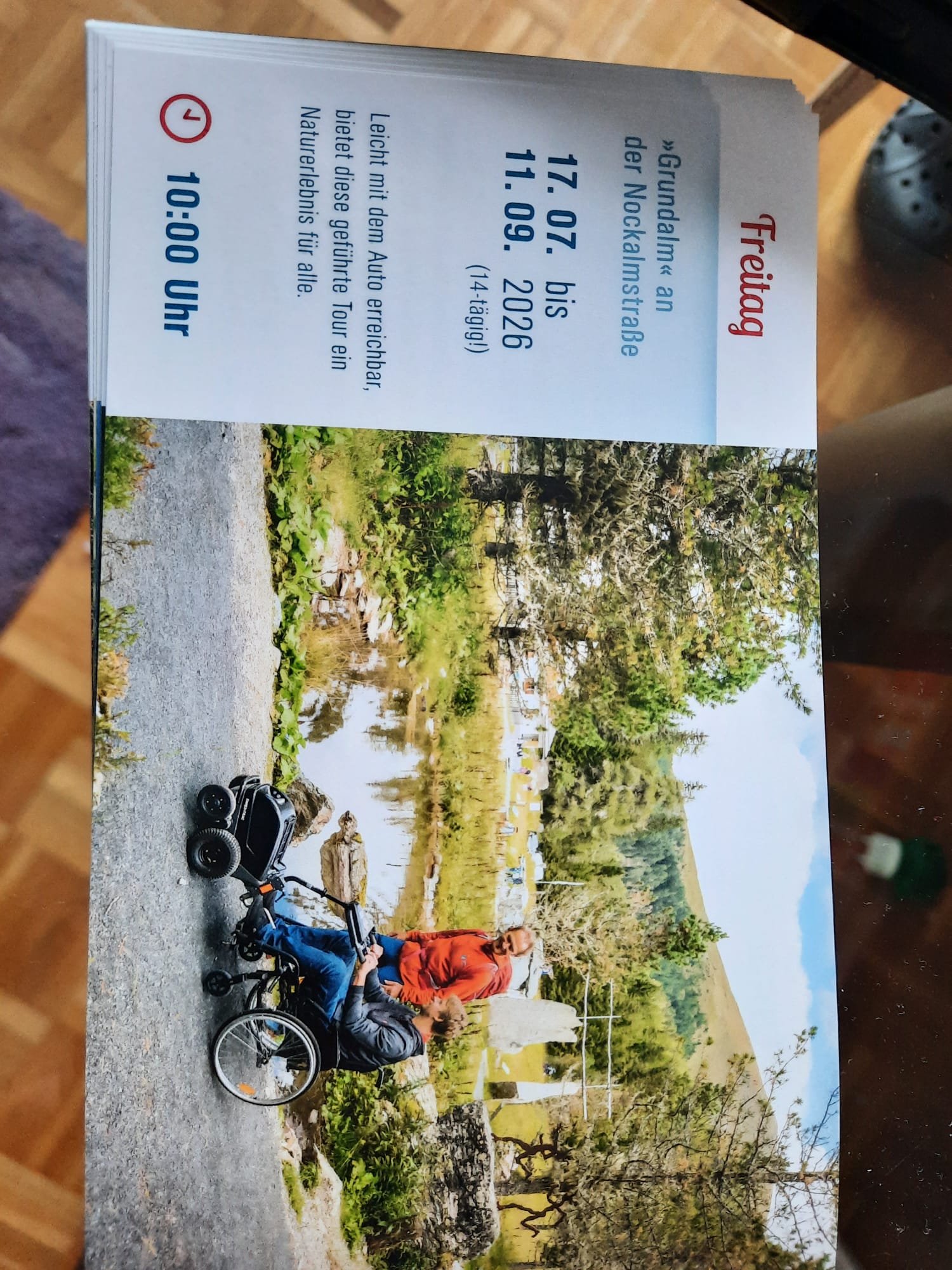 Flyer für eine barrierefreie, geführte Tour 'Grundalm' an der Nockalmstraße. Die Tour findet vom 17. Juli bis 11. September 2026 jeweils freitags um 10:00 Uhr statt. Das Bild zeigt eine Person im Rollstuhl auf einem Naturweg mit Begleitung in einer idyllischen Landschaft.